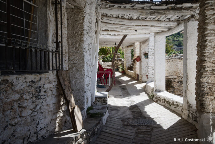 A rustic shaded walkway A sunlit, narrow stone walkway with whitewashed walls and wooden beams curves past potted plants and a red chair, leading to a view of greenery outside.