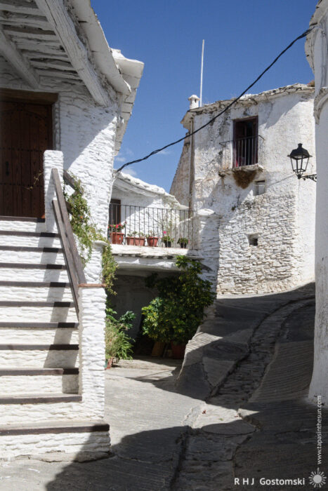 Steep stone steps to the front door A narrow, winding street in Capileira, lined with whitewashed buildings, stone steps to an entrance, potted plants, and a balcony under a clear blue sky.