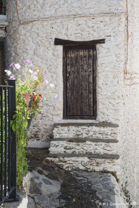 Rustic door A rustic wooden door set in a white stone wall, with uneven stone steps leading up to it and flowering plants in pots nearby. Sunlight casts soft shadows, creating a peaceful, old-world atmosphere.