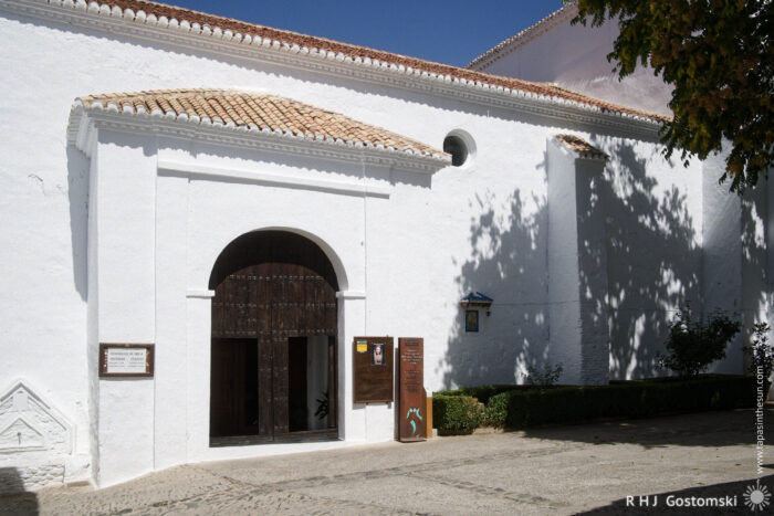 Iglesia Nuestra Seniora de la Cabeza Iglesia Nuestra Señora de la Cabeza - a white stucco building with a tiled roof and a large arched wooden entrance door, partially open. Information signs and a fuente flank the doorway. Trees cast shadows on the façade.