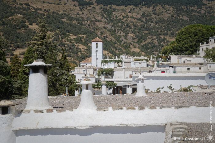 The rooftops of Capileira with the Iglesia Nuestra Seniora de la Cabeza bell tower prominent. A view of Capileira, a whitewashed village with flat roofs and distinctive Alpujarran chimneys, surrounded by green hills. A tall church tower with a tiled roof rises among the buildings.