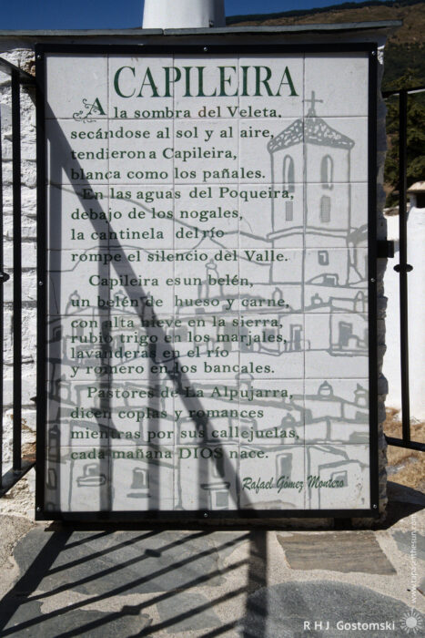 An ode to Capileira An ode about Capileira at the Mirador with Spanish text describing the village, its landscape, and traditions. The sign features green lettering, a drawing of local buildings, and is placed against a sunny outdoor backdrop with a metal railing fence.