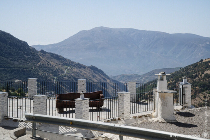 A Mirador with a view of the Poqueira Valley A brown bench sits on a terrace with white pillars and railings—this Mirador offers a stunning view of the Poqueira Valley, deep green below and framed by tall mountains under a clear sky.