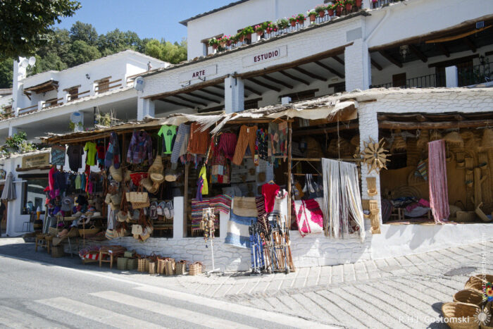 A souvenir and craft shop A vibrant souvenir and craft shop with colourful textiles, hats, bags, and crafts displayed outside a white building labelled Arte Estudio, set on a sunny day with flowers on balconies above.