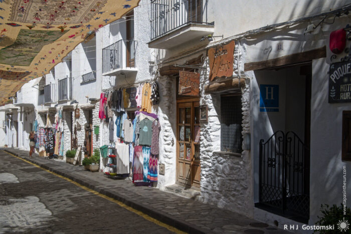 Souvenir and craft shop A narrow cobbled street lined with whitewashed buildings, colourful clothing and textiles hanging outside shops, and patterned rugs above the street providing shade. Sunlight casts shadows on the street.