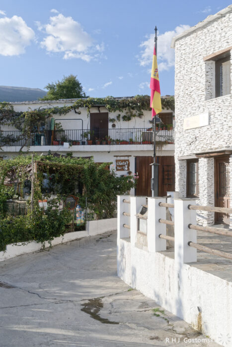 Capileira A white stone building with balconies covered in greenery and potted plants, a Spanish flag on a pole, and a clear blue sky with scattered clouds in the background.