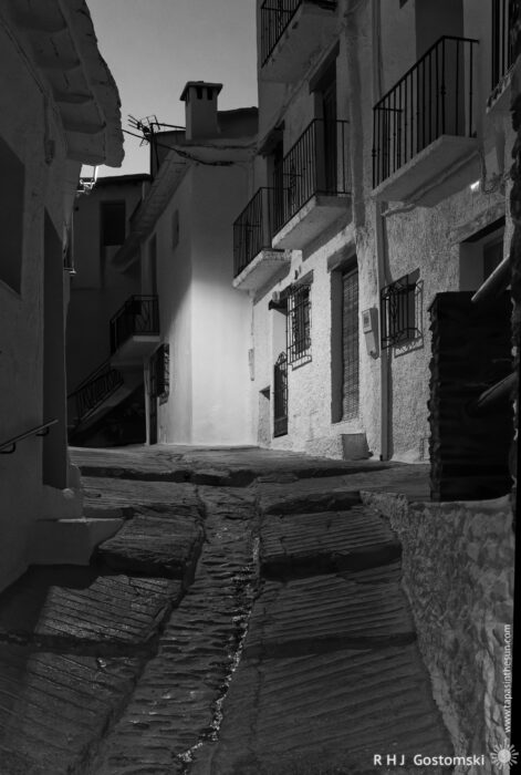 Narrow stepped alley in Capileira at night Narrow, sloped cobblestone street lined with whitewashed buildings at night; balconies and windows cast distinct shadows in the soft street lighting.