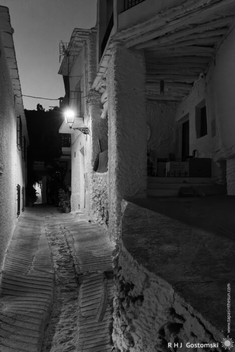 Narrow cobblestone alleyway at night Narrow cobblestone alleyway at night with whitewashed buildings, lit by a single old-fashioned streetlamp. Shadows and textures are highlighted in the black and white scene.