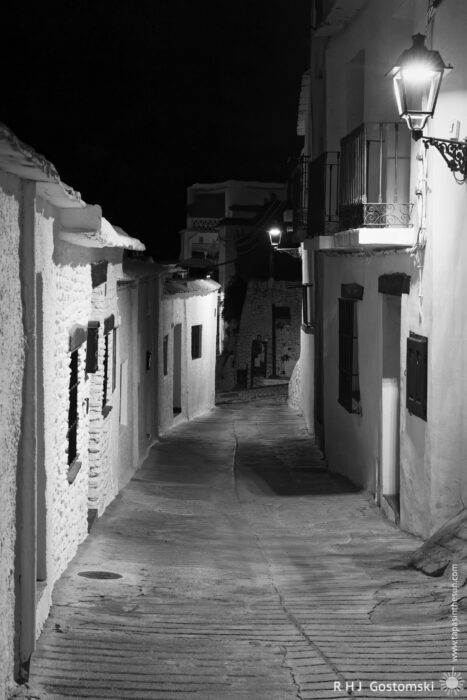 A narrow, winding street in Capileira village at night A narrow, winding street in Capileira village at night, illuminated by wall-mounted lanterns. The scene is in black and white, with whitewashed buildings and cobbled pavement.
