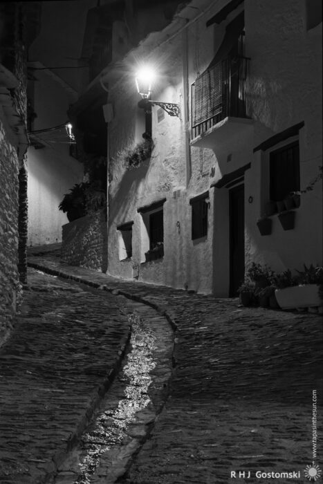 A narrow, cobbled street with central gulley A narrow, cobbled street curves uphill between whitewashed buildings at night, illuminated by a single streetlamp. The scene is quiet and empty, with shadows and reflections on the wet gulley.