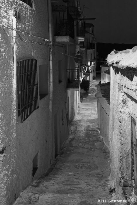 A narrow, cobblestone alleyway in Capileira A narrow, cobblestone alleyway in Capileira is lined with old buildings, lit dimly at night. Shadows and textures highlight the rough surfaces, whilst the path leads into the distance.