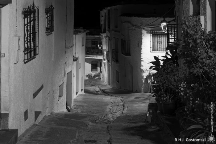 A narrow, empty street in Capileira A narrow, empty street in a village at night, illuminated by a single lamp. Whitewashed buildings line the street, with potted plants and barred windows visible. The scene is peaceful and quiet.