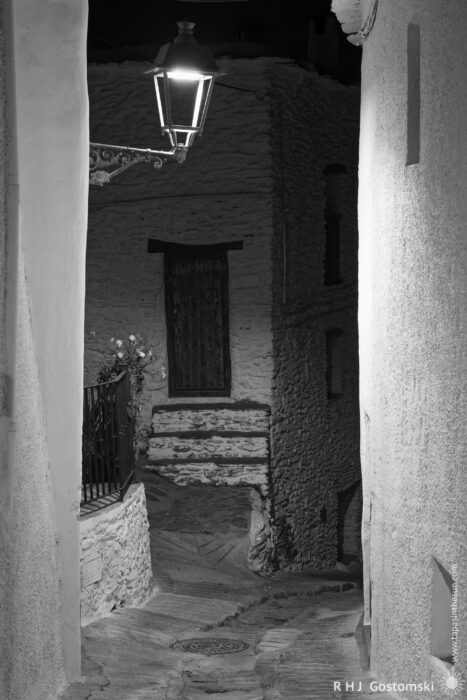 Steep steps to the front door of a village house at night A narrow stone alley at night is illuminated by a lantern. Old textured buildings and a closed wooden door are visible in the background. The scene is in black and white, creating a quiet, moody atmosphere.