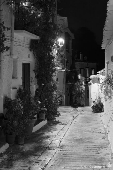 A narrow street in Capileira at night A narrow cobblestone street in Capileira, lined with white buildings and potted plants, is softly illuminated by street lamps at night. The quiet scene features shadows and climbing vines tracing the peaceful village walls.