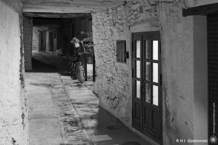 Under the tinao A narrow, cobbled alley with a tinao overhead, lined with rustic stone buildings, wooden doors, and potted plants. Bicycles are parked along one wall. The scene is in black and white, creating a nostalgic atmosphere.