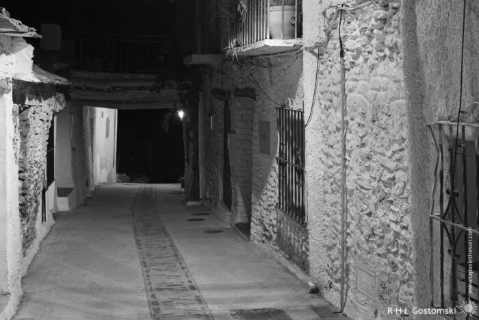 Tinao over a narrow street A narrow, empty alleyway at night, lined with old stone buildings. A single streetlamp in the distance casts a soft light, creating deep shadows on the textured walls. The scene is quiet and atmospheric.