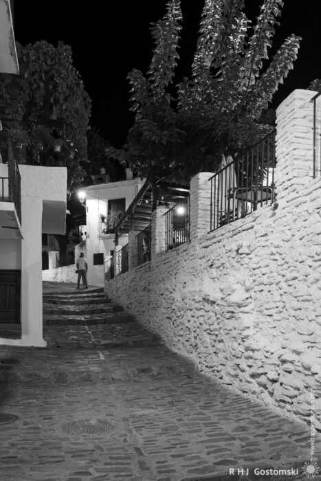 A stepped alley in Capileira A cobblestone street lined with white stone walls and outdoor lights leads uphill at night. A person stands in the distance near a tree and iron railings. The scene is quiet and illuminated in black and white.