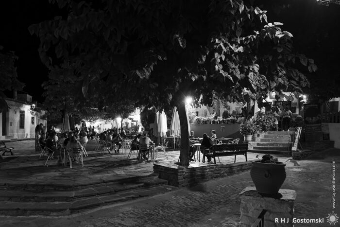 The village square at night A nighttime scene in a village square shows people sitting at outdoor tables under large trees, softly lit by surrounding lamps; buildings and stairs are visible in the background. The image is in black and white.