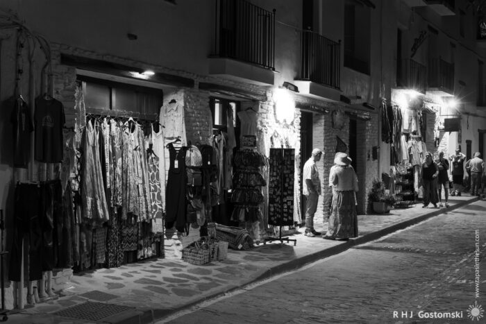 Souvenier and craft shops in Capileira at night A black-and-white photo of a narrow street at night in Capileira, where clothing, souvenir, and craft shops glow with light. Mannequins wear dresses outside, while two people, one in a hat, stand talking on the cobblestone pavement.
