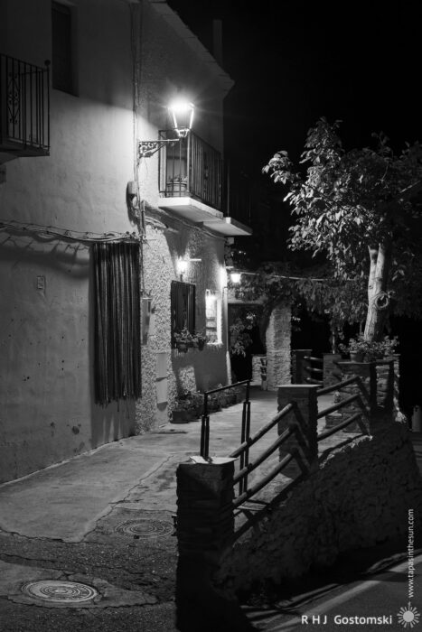 White village house in Capileira at night A quiet, narrow street at night is lit by a single wall-mounted lantern. Shadows fall on a white rendered building with balconies and shuttered windows. Trees and a stone railing line the pavement.