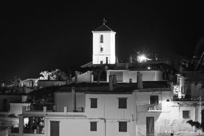 Capileira at night Black and white photo of Capileira at night, featuring white houses with tiled roofs and Alpujarran chimneys. A tall, illuminated church tower stands prominently in the background against the dark sky.
