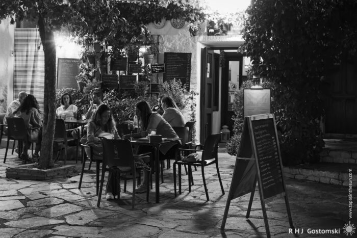 Night-life A black-and-white photo of people sitting at outdoor tables of a cosy, vine-covered café at night, softly lit by lamps, with a chalkboard menu near the entrance and stone pavement underfoot.