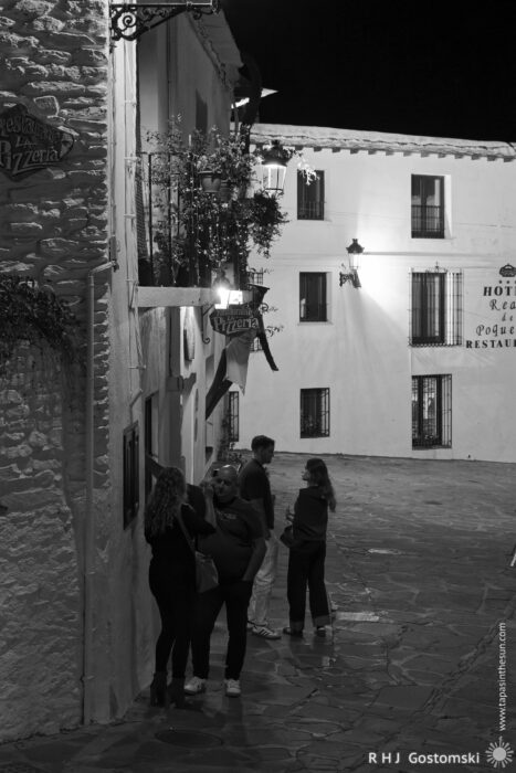 Standing and talking at night A group of people stands talking on a stone-paved street at night, beside a restaurant in the historic village of Capileira with white buildings and wrought-iron balconies. Street lamps light the scene.