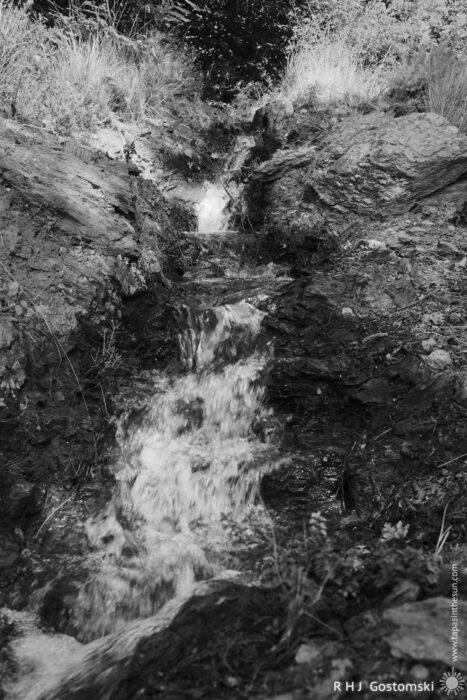 Acequia Flowing Through Rocky Terrain A small acequia cascades over rocky terrain, flowing through a natural landscape with patches of greenery and soil. Sunlight filters through surrounding vegetation, highlighting the sparkling water as it moves down the rocky path.