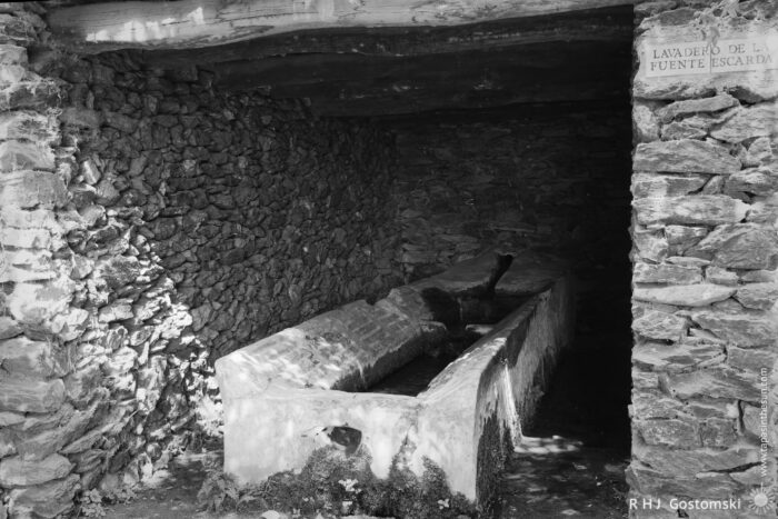 A Historic Lavadero in a Rustic Shelter A lavadero with a stone washing area with a weathered, concrete basin is enclosed by rustic stone walls. The scene is illuminated by natural light, showcasing moss and plants growing around the basin, evoking a tranquil, historical setting. A sign indicates its name.