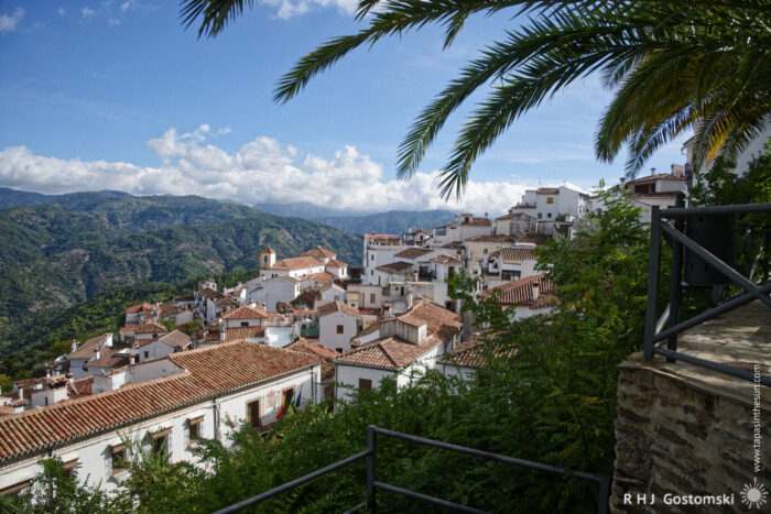 Charming White Village Overlooking Scenic Mountains A picturesque view of Benelauría nestled in the mountains, with terracotta roofs, surrounded by lush greenery and palm trees, under a bright blue sky dotted with clouds.