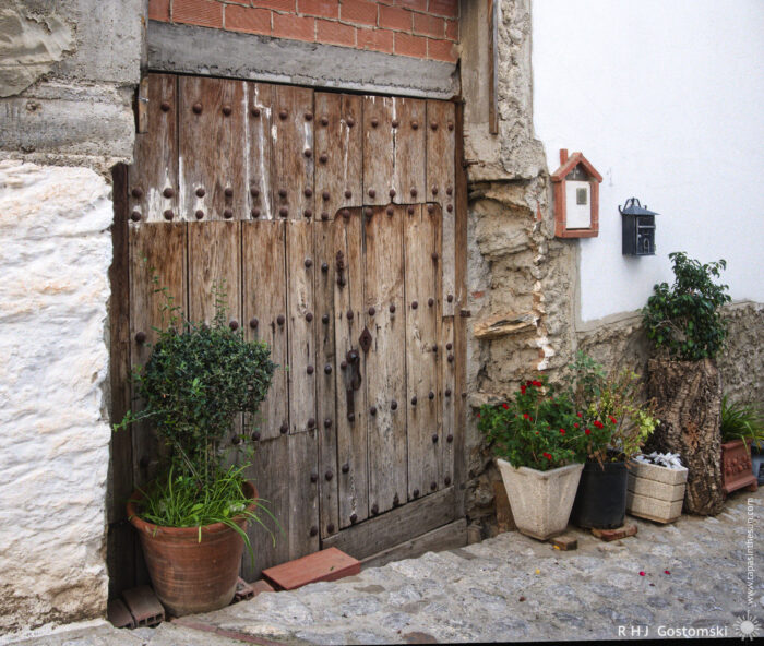 Rustic Wooden Entrance with Potted Plants A rustic wooden door set into an old stone wall, surrounded by potted plants and greenery, with a charming mailbox attached nearby. The scene captures a quaint, inviting entrance on a cobblestone path.