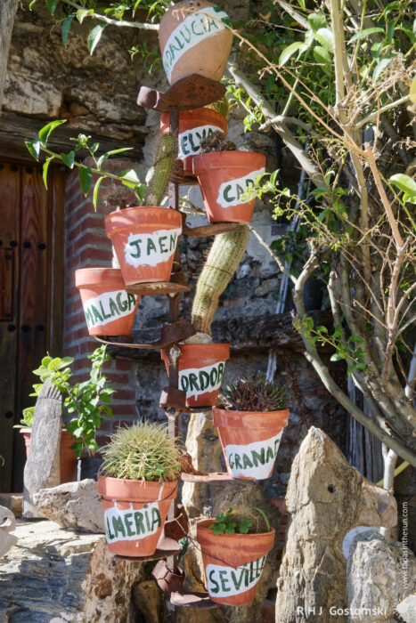 Pots Labeled With The Names of Various Spanish Cities Displayed on a Rustic Vertical Stand A vertical display of terracotta pots stacked on a rustic stand, each labeled with the names of various Spanish cities. The arrangement features cacti and greenery, set against a stone wall, creating a charming garden aesthetic.