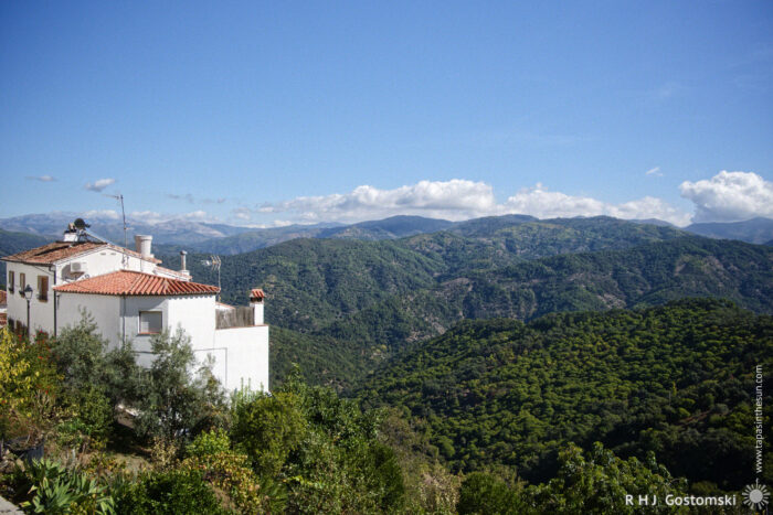 Scenic Mountain View with Rustic House A scenic view of rolling green hills and mountains under a blue sky, featuring a white house with a red-tiled roof in the foreground, surrounded by lush vegetation.