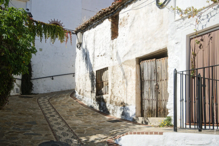 Charming cobblestone alley in Benelauría A narrow, sunlit street with whitewashed buildings featuring wooden doors, stone pavement, and a decorative pathway. Green vines drape over the wall, enhancing the charming, rustic atmosphere typical of a Mediterranean village.