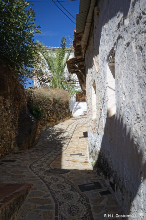 Charming Narrow Street in a Benelauría A narrow cobblestone street winds between whitewashed buildings, lined with greenery. Sunlight filters through, casting shadows and highlighting the rustic charm of the path.