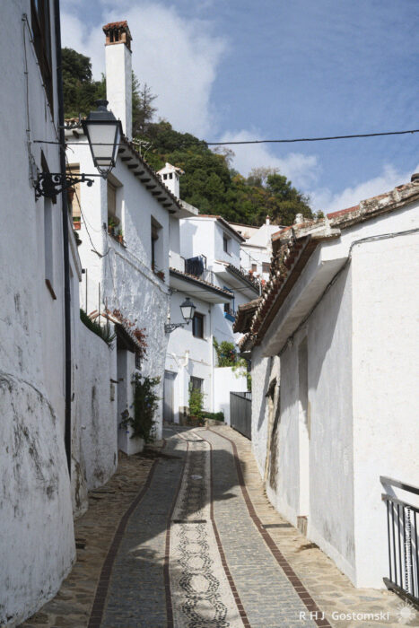 Charming Cobblestone Alleyway in the Whitewashed Village of Benelauría A narrow, winding street lined with whitewashed buildings features intricate stone paving and lush greenery, under a partly cloudy sky. The scene captures the charm of a quaint, picturesque neighborhood.