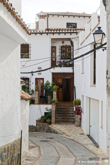 Charming Narrow Street in Benelauría A narrow cobblestone alleyway leads to a charming white building adorned with plants. The two-story structure features wooden doors and wrought iron details, creating an inviting atmosphere in a picturesque setting.