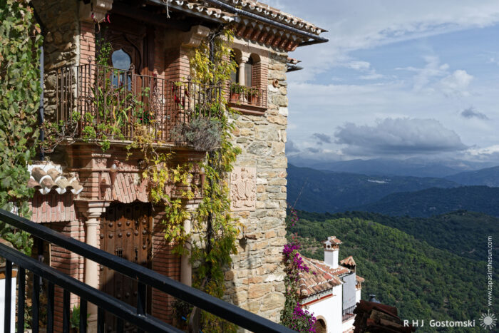 A stunning view of a traditional stone house with a wooden balcony adorned with vibrant greenery and flowers, overlooking lush green mountains and a cloudy sky.