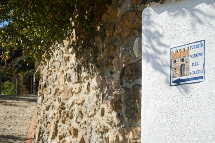 Start of the footpath to the Torreón Virgen del Rosario A weathered stone wall with a white section featuring a decorative sign that reads "TORREÓN VIRGEN DEL ROSARIO." The surroundings include green foliage and a pathway leading to the Torreón Virgen del Rosario.