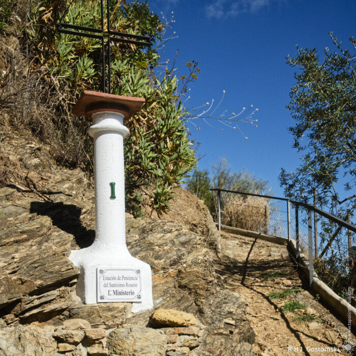 A white stone marker with a cross representing the first mystery of the rosary stands on a rocky path surrounded by greenery, with a wooden railing leading up a hillside to the Torreón Virgen del Rosario. Bright blue sky serves as a backdrop, enhancing the scenic outdoor setting.