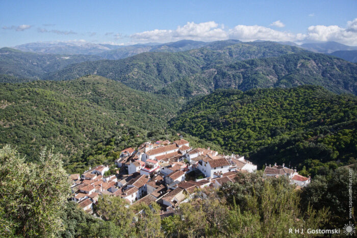 Aerial view of the picturesque white village of Benelauría nestled in a mountainous, green landscape under a clear blue sky, showcasing traditional architecture among lush forests and rolling hills.