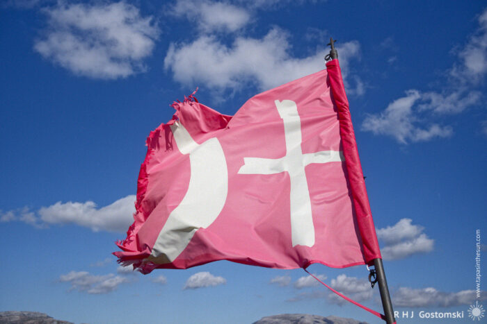 Worn Red Flag with a White Crescent Moon and Cross Against a Blue Sky A worn red flag with a white symbol resembling a crescent and a cross fluttering against a blue sky with clouds.