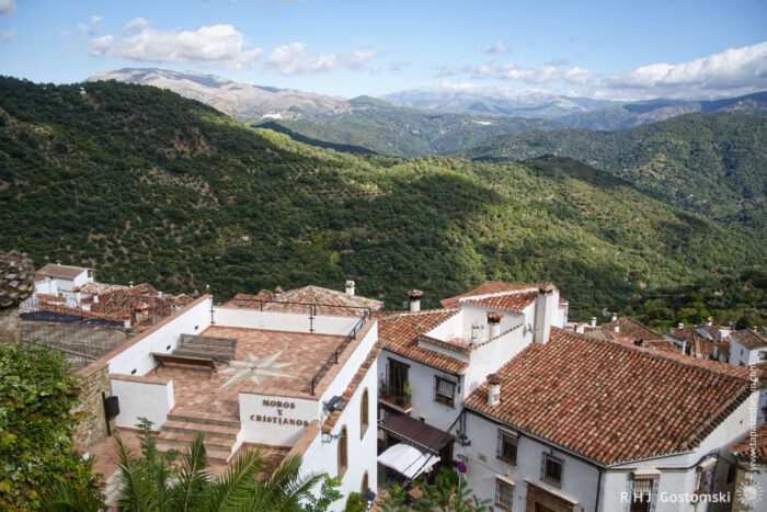 Moros and Christianos Mirador Overlooking the Village and Lush Hills Moros and Christianos Mirador overlooking a scenic view of whitewashed buildings with terracotta roofs nestled in a mountainous landscape, surrounded by lush greenery and a clear blue sky with scattered clouds.