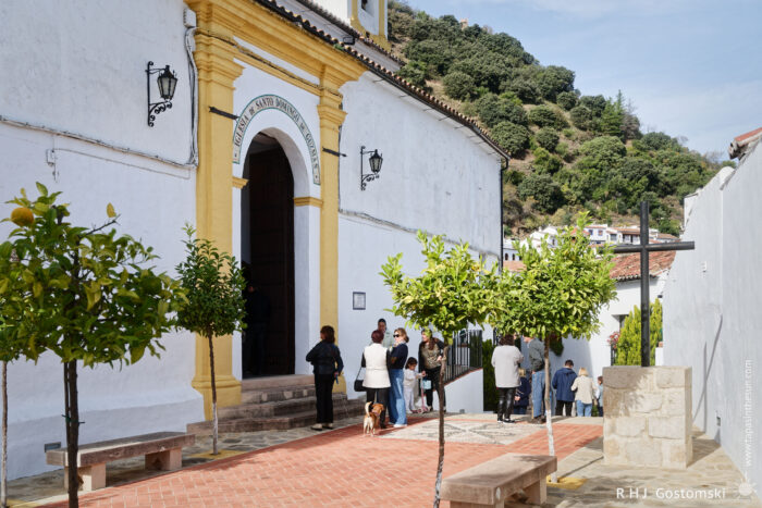 Santo Domingo de Guzmán church with yellow accents stands in a picturesque setting, surrounded by green trees. A small group of people gathers outside, enjoying the sunny day in a charming cobblestone courtyard, with hills in the background.