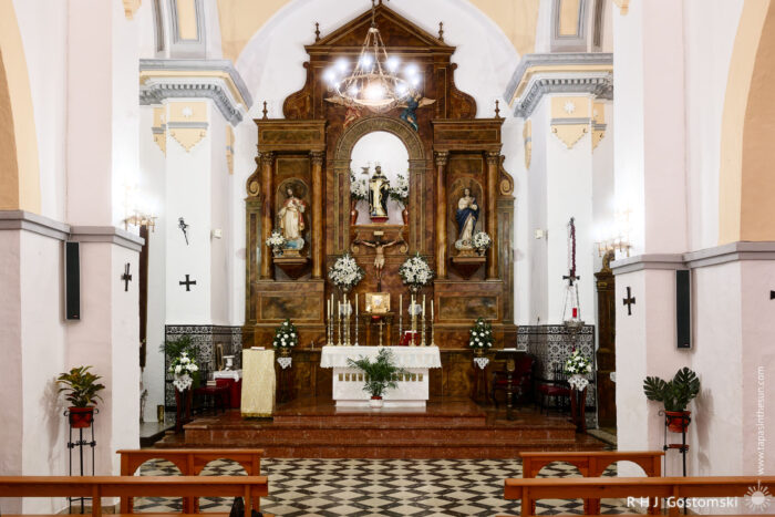 Interior of Santo Domingo de Guzmán church featuring a decorative altar with religious figures, surrounded by flower arrangements. The space includes wooden benches, a checkered floor, and soft lighting, creating a peaceful and reverent atmosphere.
