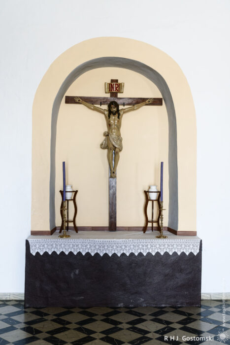 Crucifixion Altar in a Minimalist Church Setting A wooden crucifix of Christ is mounted on a wall above a simple altar, flanked by two candlesticks, with a white cloth draping the front. The altar is set against a neutral-toned background, featuring a checkered black and white floor.