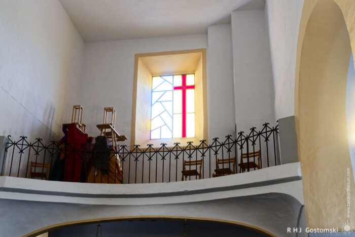 A view of the choir balcony inside Santo Domingo de Guzmán church interior featuring a colorful stained glass window, a railing with decorative elements, and wooden chairs arranged on a balcony. The space is bright and serene, characterized by simple architectural lines and minimal furnishings.