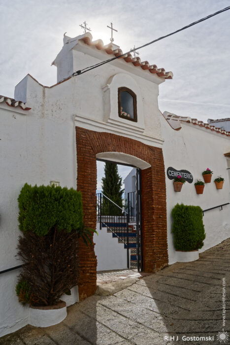 Aa archway leads to a cemetery entrance, framed by greenery and flower pots, under a bright sky with a cross at the top. The scene conveys a peaceful yet somber atmosphere.