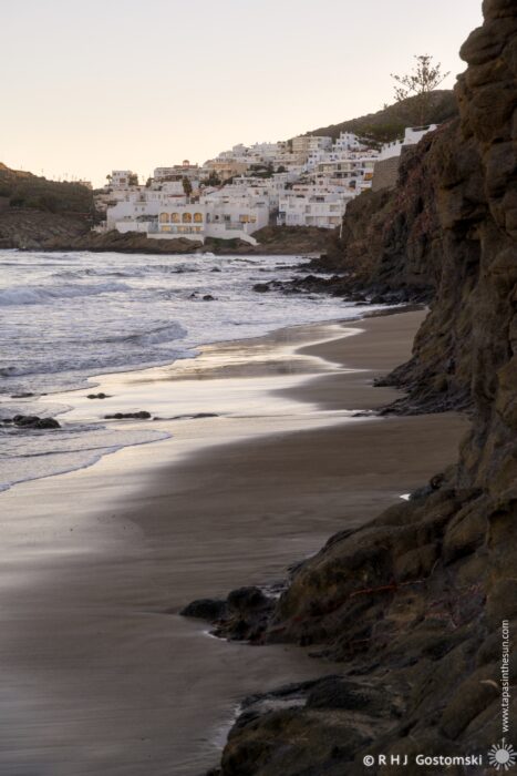 A serene beach scene at dusk, featuring gentle waves lapping against a sandy shore. In the background, white buildings are nestled among the cliffs, creating a picturesque coastal view.