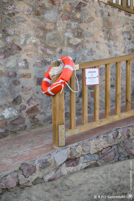 A bright orange lifebuoy attached to a wooden railing beside a stone wall, with a warning sign nearby, indicating safety precautions.